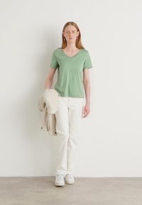 Grønn V-hals t-skjorte, mykt materiale, kombinert med hvite straight-leg jeans og hvite sneakers. Jakke lagt over venstre arm.