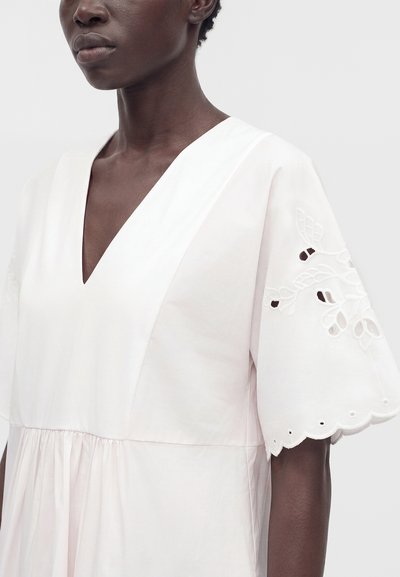 Femme portant une robe blanche avec un décolleté en V profond et des manches larges ornées de motifs floraux découpés et brodés, ainsi que des bords festonnés.