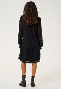 Vestido negro de encaje con mangas largas y un dobladillo en capas, con un patrón floral y tejido texturizado. Combinado con botas negras hasta el tobillo.