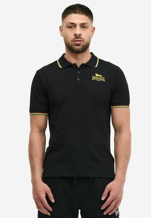 Uomo che indossa una polo nera Lonsdale con rifiniture rosse, gialle e verdi su colletto e maniche, in piedi davanti a uno sfondo bianco uniforme.