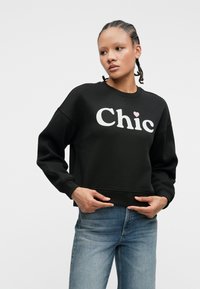 Woman with braided hair wearing a black sweatshirt with "Chic" and a pink heart, paired with light blue jeans, posing against a white background.