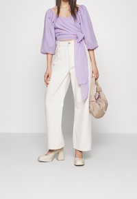 Ljuslila omslagstopp med puffärmar, vita vida byxor och beiga högklackade skor. Beigefärgad väska med knuthandtag.