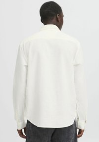 White long-sleeved shirt with a collar, buttoned cuffs, and a loose fit. Smooth texture with a subtle sheen, hem curved at the sides.