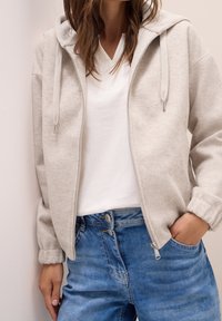 Jasnoszara bluza na zamek z teksturą polarową i regulowanymi sznurkami, noszona na białej koszulce i zestawiona z niebieskimi dżinsami.
