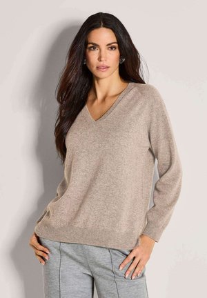 Een vrouw met lang, donker haar, gekleed in een beige V-hals trui en grijze broek, staand met haar handen op haar heupen tegen een lichte achtergrond.