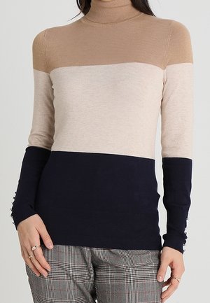 Femme portant un pull à col roulé color-block beige, crème clair et bleu marine, associé à un pantalon gris à carreaux.