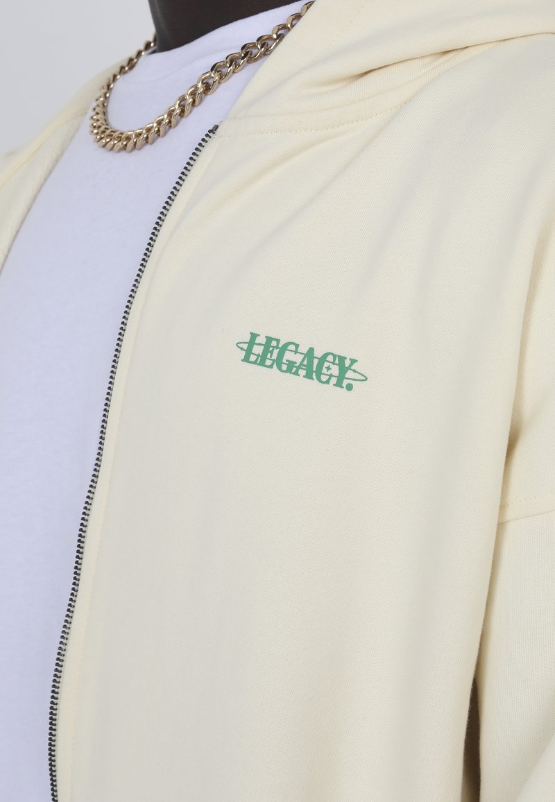 Sudadera crema con un logotipo verde "LEGACY", cremallera frontal y tejido texturizado. Usada sobre una camiseta blanca con una cadena dorada.