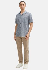 Hellblaues kurzärmliges Hemd mit Knöpfen und einer Tasche, kombiniert mit hellbraunen Hosen und weißen Sneakern. Das Modell steht vor einem schlichten Hintergrund.