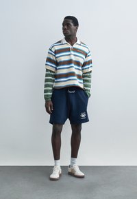 Hombre con camiseta de manga larga de rayas en capas, pantalones cortos azules marinos con parche de logotipo, calcetines blancos y zapatillas crema de pie sobre un suelo gris.