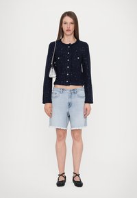 Marinefarget teksturert cardigan med knapper og sølvdetaljer, sammen med lyseblå denimshorts med frynser og svarte prikkete leiligheter.