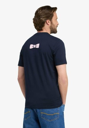 Homme aux cheveux bruns et à la barbe portant un t-shirt bleu marine avec un motif de nœud rose dans le haut du dos et un jean bleu.