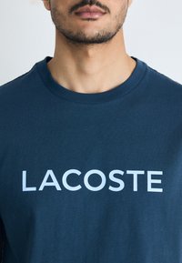 Tamnoplava pamučna majica s bijelim "LACOSTE" logotipom sprijeda; okrugli izrez, kratki rukavi i glatka tekstura.