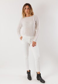 Donna in piedi con le mani nelle tasche di pantaloni bianchi, indossando un maglione lavorato a maglia beige chiaro e stivaletti neri alla caviglia su uno sfondo semplice.