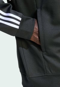 Μαύρη αθλητική κουκούλα με λευκό σχέδιο τριών λωρίδων στα μανίκια, που διαθέτει υφή υφάσματος, πλαινές τσέπες και  ribbed μανσέτες.