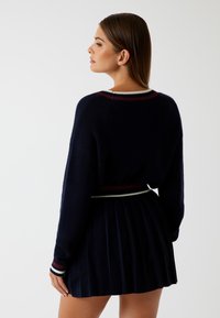 Vrouw die naar links kijkt, gekleed in een donkerblauw geplooide rok en een bijpassende gebreide trui met rode en witte gestreepte rand.