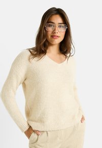 Pull crème à col en V en tissu doux et texturé. Coupe décontractée, manches longues et ourlet côtelé. Associé à un pantalon clair à rayures.