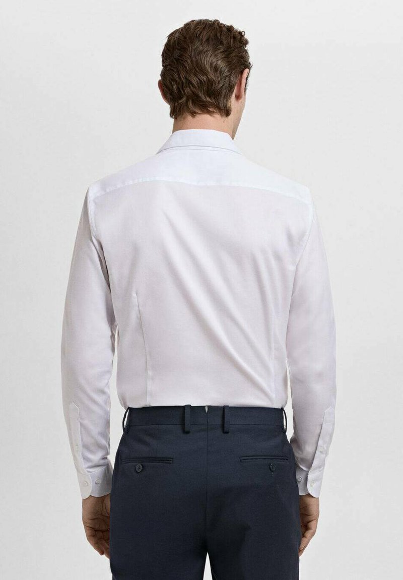 Chemise blanche à manches longues avec une texture lisse, rehaussée d’un empiècement discret aux épaules et de boutons aux poignets, associée à un pantalon foncé.
