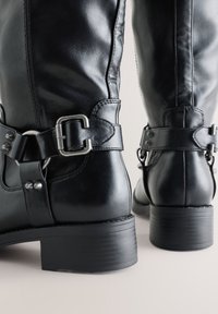 Schwarze Leder-Mittelkalbsstiefel mit silbernen Schnallenriemen, runden gestapelten Absätzen und glatter strukturierter Oberfläche, von hinten auf einer hellen Oberfläche gezeigt.