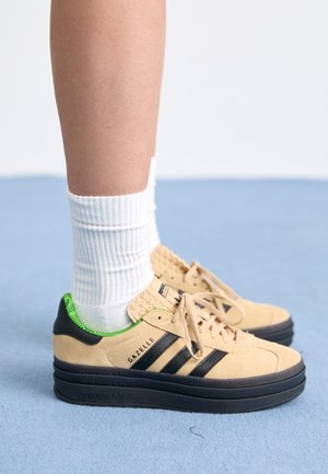 Nogi w białych prążkowanych skarpetkach i beżowych trampkach Adidas Gazelle z czarnymi paskami i grubymi czarnymi podeszwami na jasnoniebieskiej powierzchni.