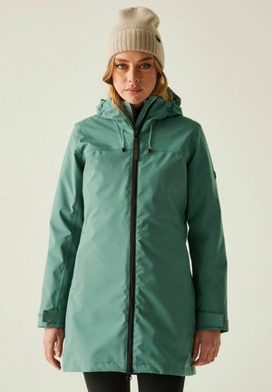 Kobieta w turkusowej wodoodpornej kurtce z kapturem i beżowej dzianej czapce, stoi na jednolitym jasnym tle, zwrócona twarzą do przodu.