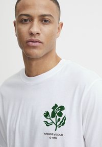 Camiseta de algodón blanca con un gráfico floral verde en el lado izquierdo del pecho, con el texto "ARCHIVE of SOLID © 1999" debajo del diseño.