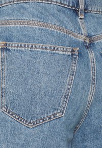 Primer plano de un par de jeans de mezclilla azul, que presentan un solo bolsillo trasero con una forma puntiaguda, costuras naranjas y un acabado de tela texturizada.