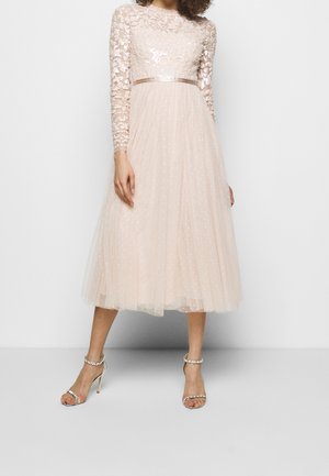 Kobieta ubrana w bladoróżową, midi sukienkę z długim rękawem, z koronkowym gorsetem ozdobionym kwiatowym motywem oraz tiulową spódnicą, w połączeniu z wysokimi sandałami ozdobionymi perłami.