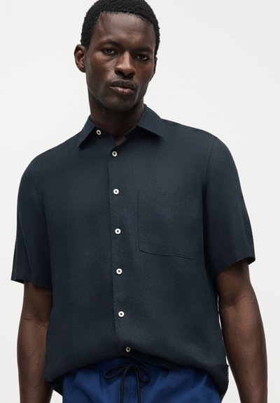 Άνδρας που φοράει σκούρο μπλε κοντομάνικο πουκάμισο με τσέπη στο στήθος και μπλε παντελόνι με κορδόνι σε απλό φόντο.