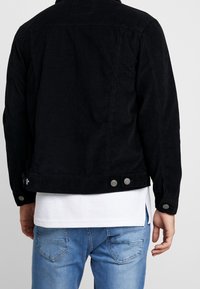 Man i svart kordjacka över en vit skjorta och ljusblå jeans, sedd bakifrån mot en enkel bakgrund.