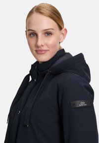Veste à capuche navy avec une texture lisse, dotée d'une fermeture éclair, de cordons ajustables et d'un patch logo sur la manche.