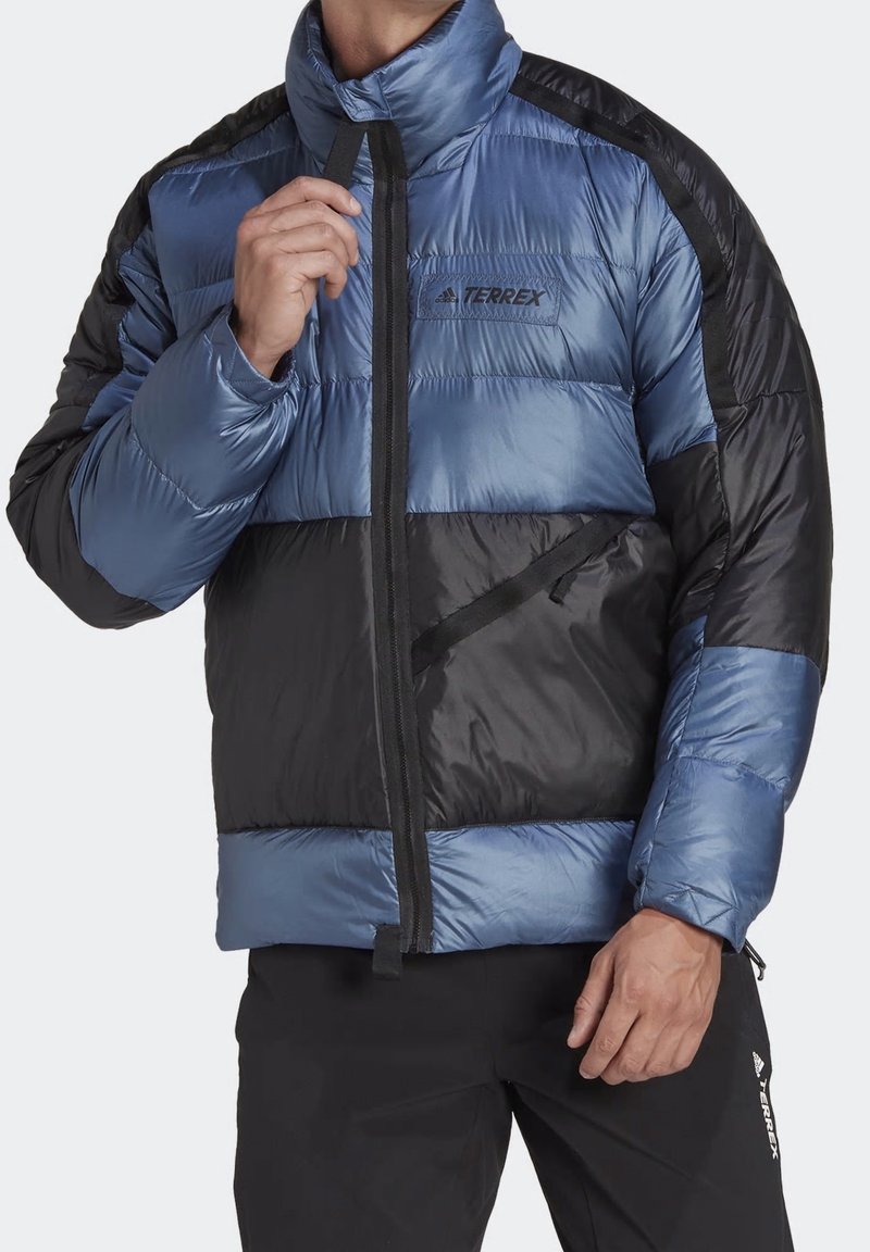 Homme portant une veste matelassée Terrex bleue et noire, fermeture éclair partiellement fermée, avec un pantalon Terrex noir.