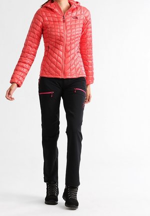 Bright coral puffer jacket with a hood, quilted texture, zip pockets; paired with black pants featuring pink zip details and black hiking boots.