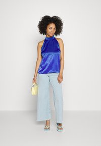 Top halter azul de cetim, jeans de perna larga azul claro, bolsa amarela e sandálias azuis com tiras. O top tem uma textura suave e um acabamento brilhante.