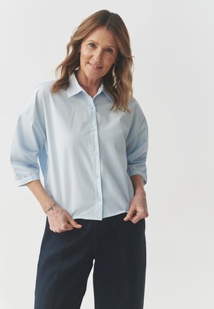 Light blue button-up shirt with a collar, three-quarter sleeves, and a relaxed fit, paired with dark pants on a neutral background.