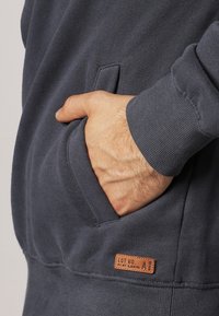 Ręka częściowo wsunięta do przedniej kieszeni ciemnoszarej bluzy z prążkowanym mankietem i małą brązową skórzaną łatą w pobliżu dolnego brzegu.