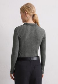 Blondynka z włosami związanymi w kucyk, ubrana w szary, prążkowany top z długim rękawem włożony w czarne spodnie z czarnym paskiem, stojąca przodem do białej ściany.