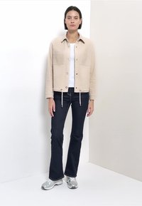 Veste beige en velours côtelé avec deux poches avant, portée sur un haut blanc, associée à un jean évasé foncé et des baskets argentées. Fond uni.