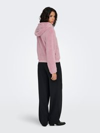 Sweat à capuche rose et duveteux avec capuche, ourlet côtelé et poches. Porté avec un pantalon noir à jambes larges et des bottes noires à talons. Fond uni.