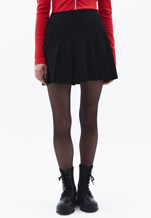 Zwarte plissé minirok met een aansluitende taille, gecombineerd met doorschijnende zwarte panty’s en chunky zwarte combatboots. Rood langemouwen top zichtbaar.
