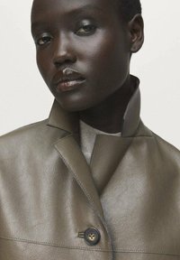 Gros plan d'une personne à la peau foncée portant une veste en cuir olive à col haut, regardant légèrement vers le bas avec une expression neutre.