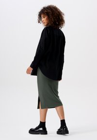 Black long-sleeve shirt, green ribbed midi skirt with side slit, and black platform shoes. Curly hair adds texture. Plain background.