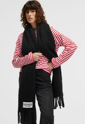 Black knitted scarf with fringed edges, made of soft material, worn over a red and white striped long-sleeve shirt, featuring a logo patch.