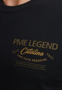 Crna majica s kratkim rukavima i okruglim izrezom s zlatnim natpisima: "PME LEGEND", "Catalina" i "FIND YOUR FREEDOM". Glatka tkanina, prilagođeni kroj, minimalni ukrasi.