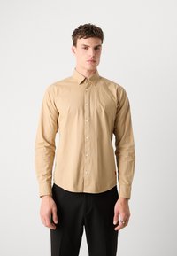 Beige long-sleeve shirt in lightweight fabric, buttoned front, pointed collar, and rounded hem. Features button cuffs and a small logo detail.