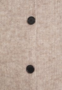 Närbild av beige stickad tyg med två svarta fyrhålsknappar vertikalt centrerade.