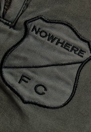 Nærbillede af en mørkegrøn stofpatch med sort broderi, hvor der står "NOWHERE" og bogstaverne "F" og "C" adskilt af en vandret linje.