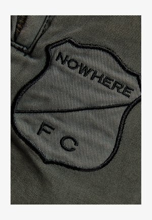 Nærbillede af en mørkegrøn stofpatch med sort broderi, hvor der står "NOWHERE" og bogstaverne "F" og "C" adskilt af en vandret linje.