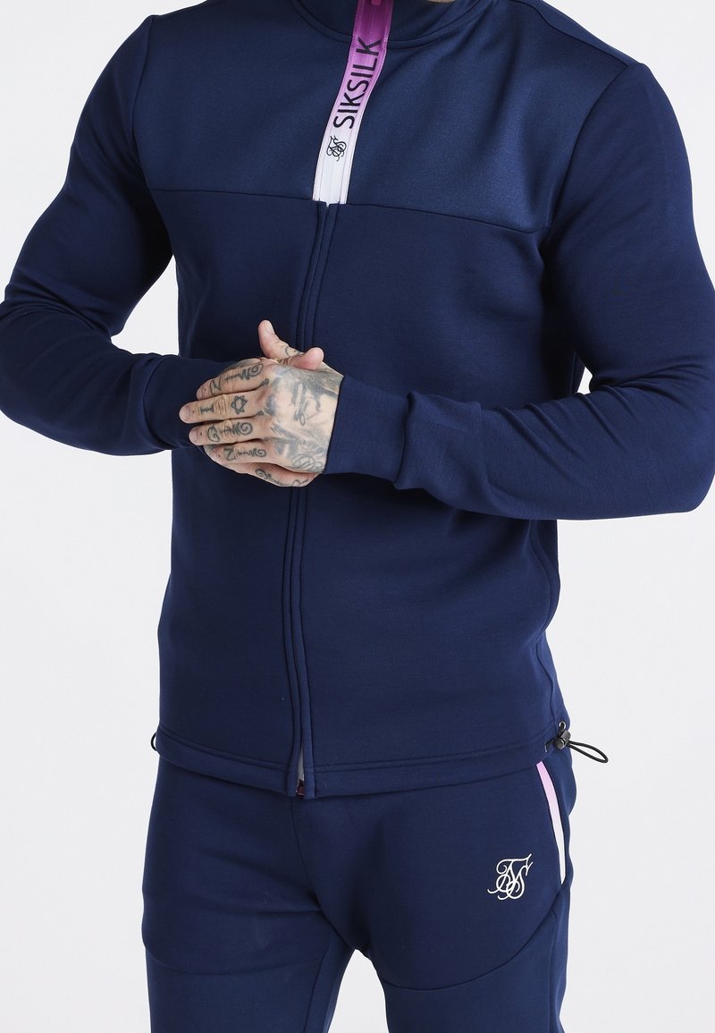 Giacca atletica della marina con chiusura a zip, dettagli a collo viola a contrasto e tessuto elasticizzato. Presenta un design aderente e accenti di logo sottili.