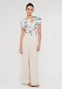 Blouse à imprimé floral blanc avec des flamants roses, associée à un pantalon large taille haute beige avec un détail de bouton.
