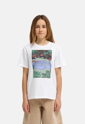 Młoda osoba z prostymi brązowymi włosami, ubrana w białą koszulkę z kolorową ilustracją basenu i otaczających roślin.